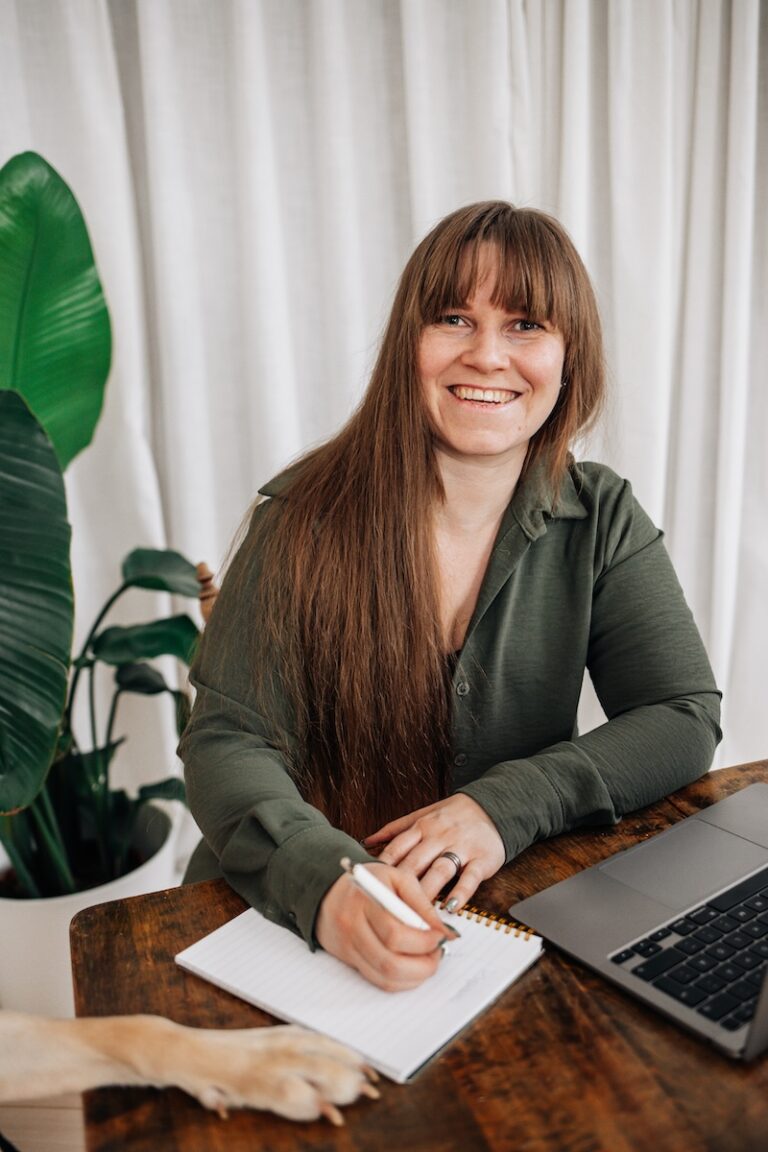 Redovisningskonsult sitter vid ett träbord och skriver i ett anteckningsblock med laptop bredvid, i ljus inomhusmiljö med grön växt i bakgrunden.